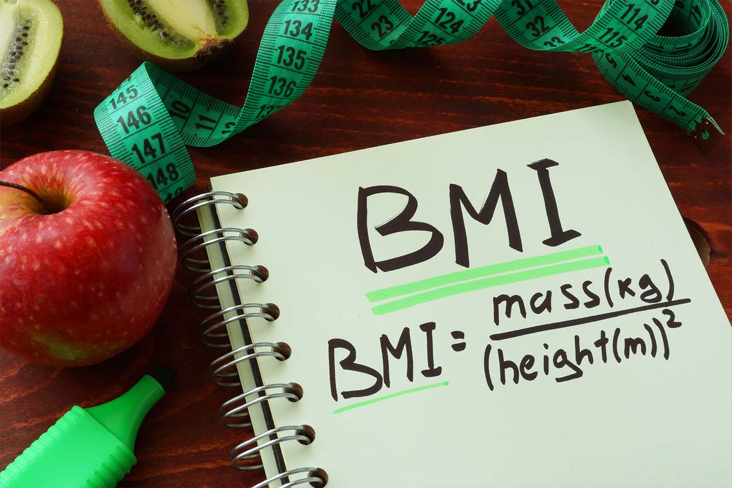 Notebook with BMI formula, apple, kiwi, and measuring tape.