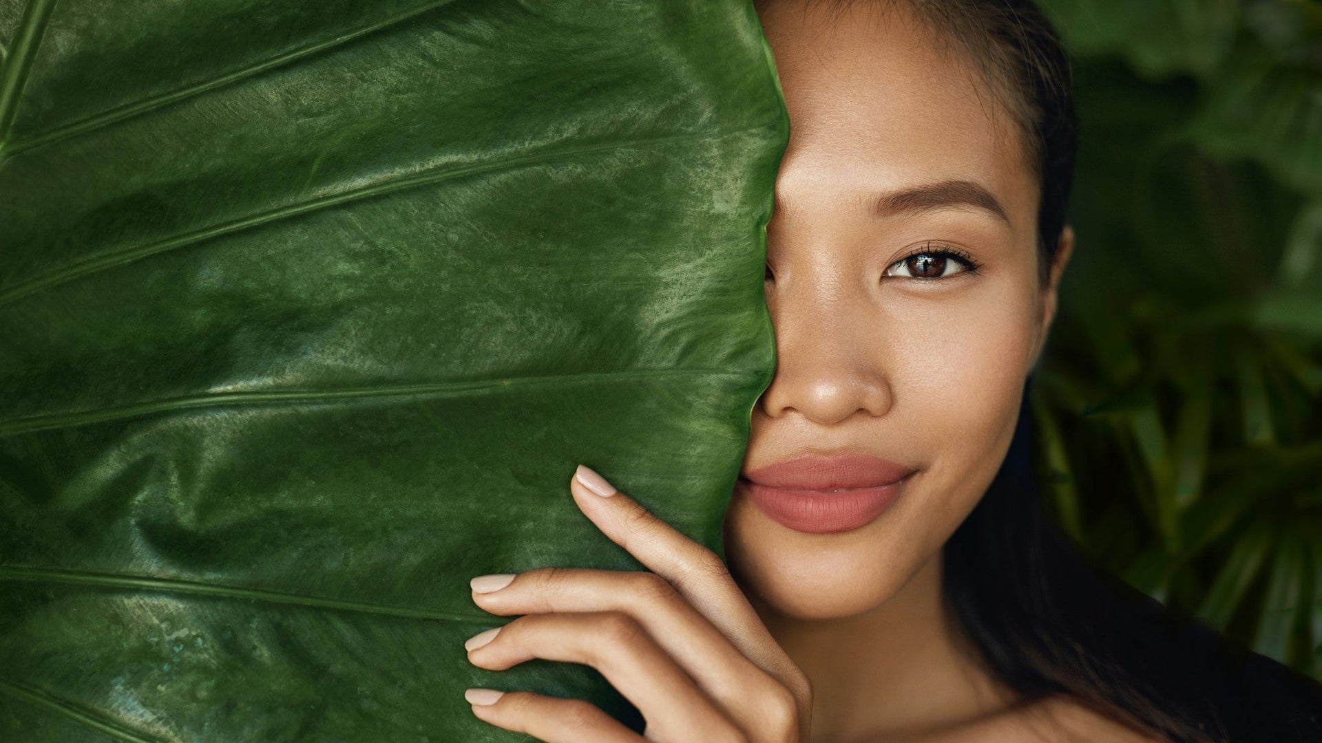 Woman with clear skin peeking from behind large green leaf.