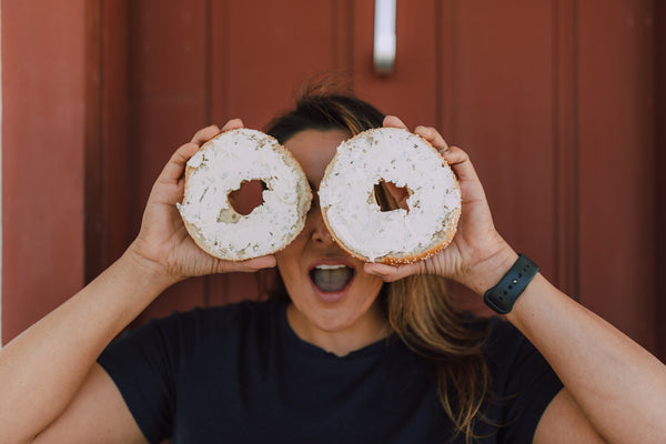Woman playfully holding two bagels with cream cheese over her eyes.