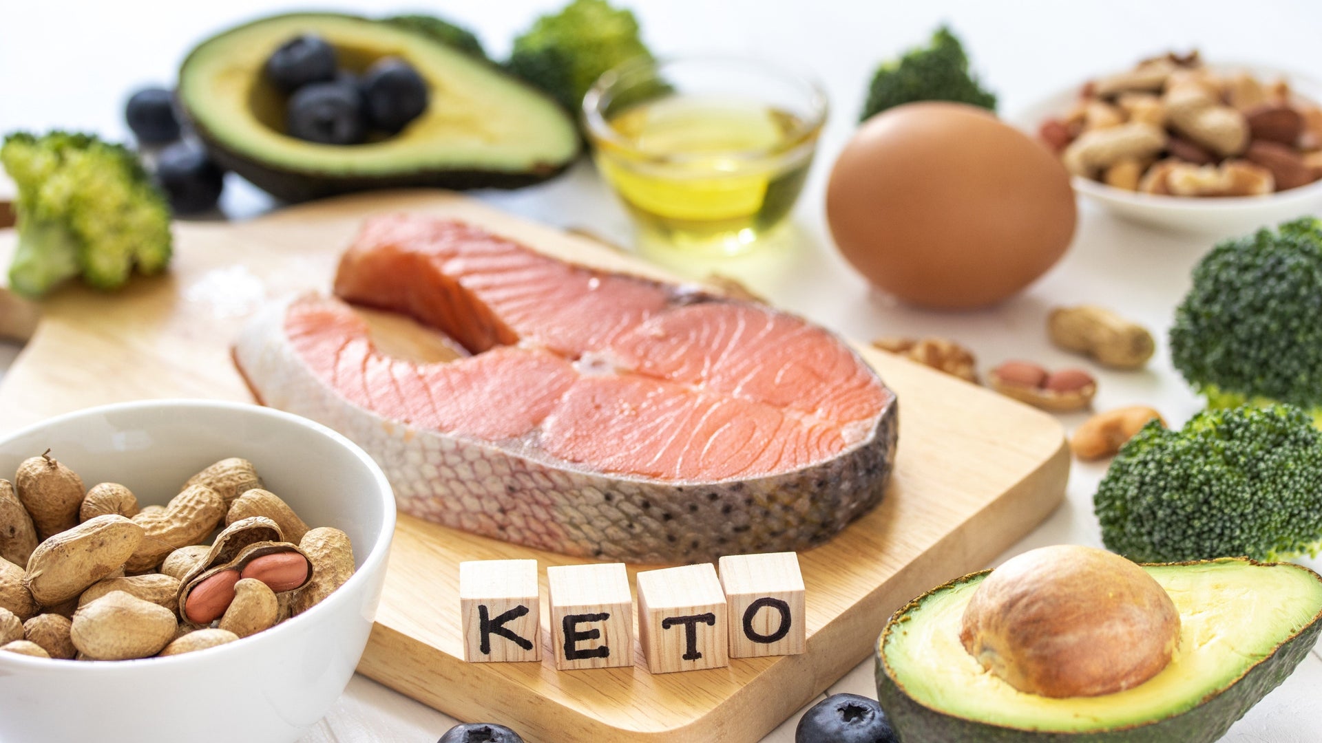 Keto-friendly foods including salmon, avocado, nuts, broccoli, and egg on a wooden board.